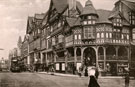 View: c12522 Postcard view of Eastgate Street viewed from the Cross. The Eastgate and Clock are visible to the left of image. Electric tram and pedestrians seen at street level. The building on the corner is 1 Bridge Street, with steps leading up to the Rows.